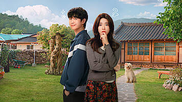 A young man and woman stand in a sunny, rural yard with traditional wooden house, lawn, garden statues, and a fluffy dog, mountains in the background, blue sky with clouds, both subjects displaying thoughtful and playful expressions.