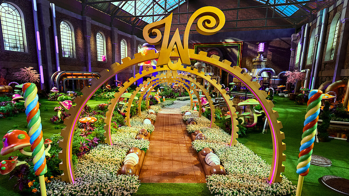 A whimsical candy-themed garden inside a large factory with colorful sweets, oversized mushrooms, lollipops, golden arches, flowers, and a large "W" centerpiece under a glass roof, creating a magical, playful atmosphere.