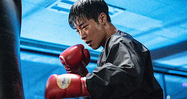 In a blue-lit room, a man wearing a black jacket and red boxing gloves prepares to hit a punching bag.
