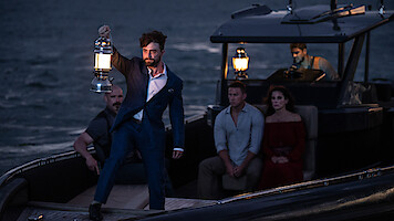 A man in a blue suit holds up a lantern on a small boat at dusk, surrounded by four other people sitting and watching, with a dark sea and another boat in the background.