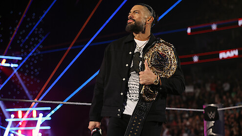 Roman Reigns holding a championship belt stands confidently in a wrestling ring, surrounded by colorful lights and a cheering crowd during a live event.