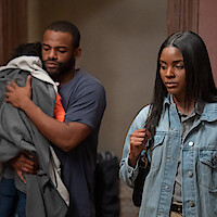 A man holding a child stands in the background while a woman with long hair, wearing a denim jacket and carrying a bag, looks serious in the foreground. They appear to be indoors, possibly in a home or apartment hallway.