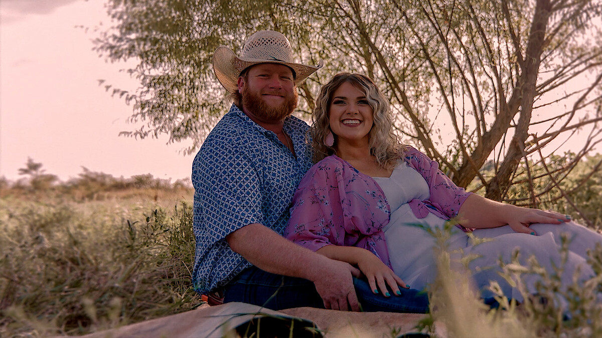Smiling couple sitting on a blanket outdoors in a grassy field, with trees in the background, wearing casual clothes and cowboy hat, enjoying a relaxed moment in nature during daylight.