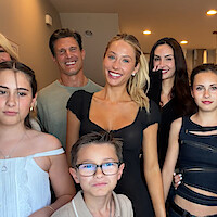 A smiling group of adults and children posing together indoors in a well-lit, modern home setting with neutral colored walls and ceiling lights.