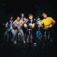 Seven teenage boys on BMX bikes pose together at night outdoors, dressed in casual, colorful streetwear. The group is illuminated, creating a dramatic, moody, and nostalgic atmosphere, suggesting friendship and adventure.