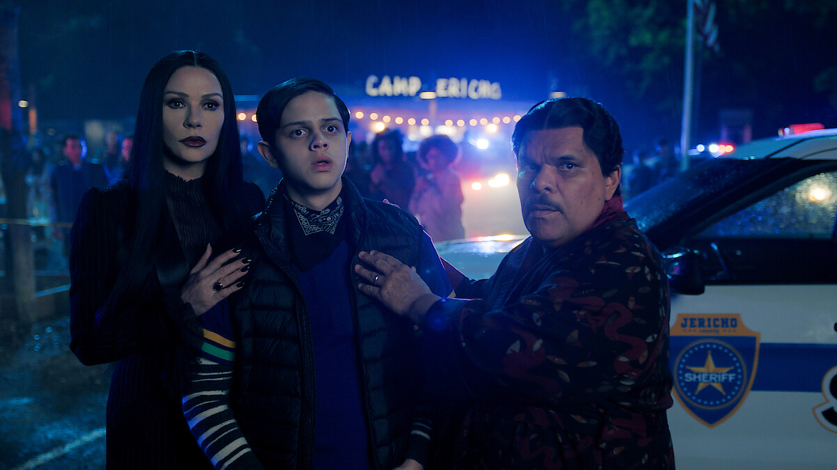 Catherine Zeta-Jones as Morticia Addams, Isaac Ordonez as Pugsley Addams, and Luis Guzmán as Gomez Addams stand in front of a police car in 'Wednesday' Season 2.