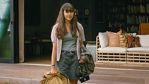 Young woman with long hair stands outside a cozy, modern house holding a coat, bag, and camera. The relaxed setting features a wicker sofa and bookshelves, evoking a calm, casual, and welcoming atmosphere.