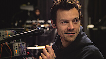 Harry Styles in a hoodie sits at a microphone with electronic equipment around him in a dimly lit studio or rehearsal space, looking off to the side and smiling slightly.