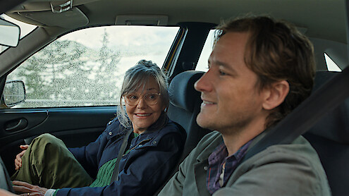 Two people sitting inside a car, one woman driving and smiling at the passenger, a man who is relaxed and smiling. The car is parked with a window showing a scenic outdoor background with trees and light rainfall.