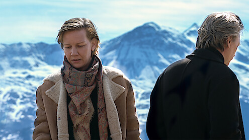 Two people stand outdoors in winter clothing with snowy mountains in the background, one facing forward and the other with their back turned, suggesting a somber or reflective mood in a cold, alpine environment.