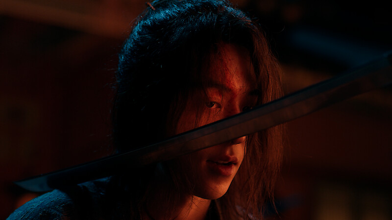 A person with long hair and a cut on their forehead is in a dimly lit, dramatic setting, looking forward with a sword held close to their face, suggesting tension or confrontation.