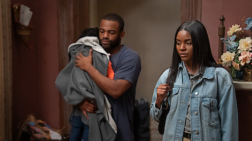 A man holding a child stands beside a woman in a denim jacket. They are indoors in a warmly lit room with flowers on a shelf and decorative objects in the background. The mood appears serious or contemplative.