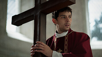 A solemn priest in red vestments holds a large wooden cross inside a softly lit church, conveying a contemplative and reverent atmosphere.