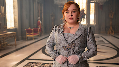 Woman in a detailed period costume stands in an ornate, sunlit palace room with chandeliers, gilded furniture, and marble floors.