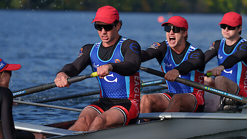Rowers in matching uniforms and red hats compete in a crew race on a calm river, focused and working together in an outdoor setting with greenery in the background.