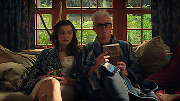Rachel Weisz as M and John Slattery as John in bathrobes sit on a cozy couch by large windows, one holding a drink, the other reading a book, surrounded by cushions and natural light, with greenery visible outside.