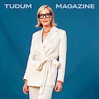A woman with blonde hair and glasses wearing an elegant white suit poses confidently in front of a blue backdrop with "Tudum Magazine" text at the top.