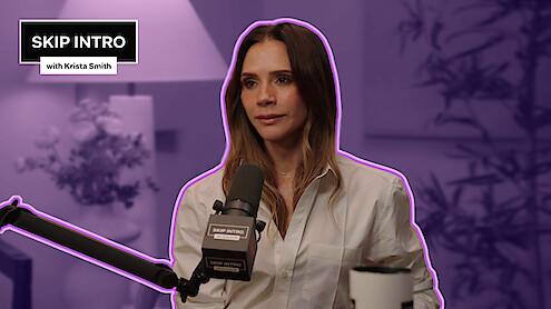 Victoria Beckham in a white shirt speaks into a microphone during a podcast interview, sitting in a softly lit studio with plants and a lamp, mood is calm and focused, overlay text reads "Skip Intro with Krista Smith".