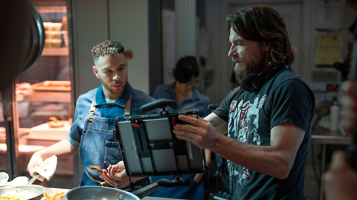 A director on set in casual dress shows an actor a tablet as they work together on a shot.