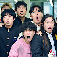 A group of diverse students, wearing casual and school uniforms, stand closely together indoors, expressing surprise and excitement, with two of them having visible bandages on their faces. Bright, modern school environment.
