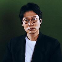 Man with glasses and dark hair wearing a black blazer over a white shirt, sitting against a plain green background in studio lighting.
