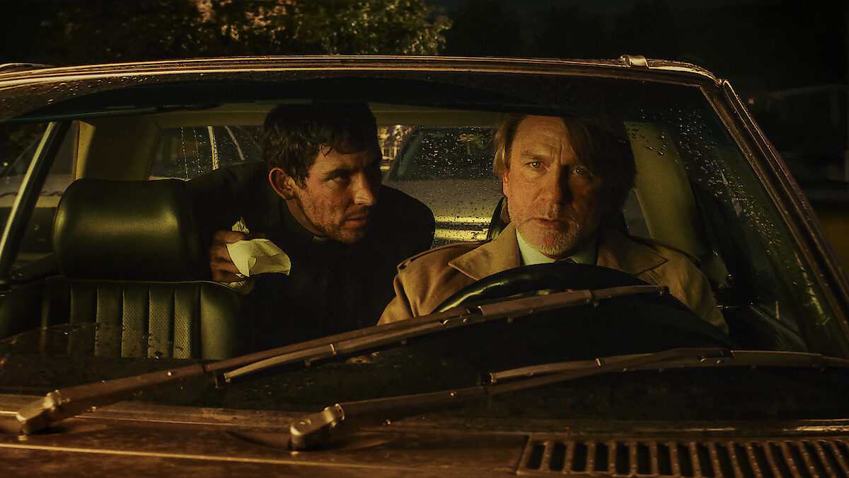Two men sit in a car at night, illuminated by dim streetlights and rain on the windshield. The driver looks concerned while the passenger leans in with urgency, creating a tense and suspenseful atmosphere.