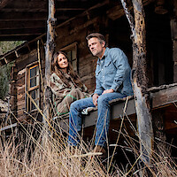 A man and woman sit on the porch of a rustic wooden cabin surrounded by tall grass, wearing casual outdoor clothing. The setting appears peaceful and rural, with natural wooden beams and a weathered structure suggesting a remote location.