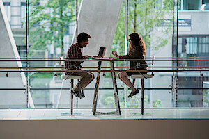 Two people sit across from each other at a high table in a modern, glass-walled building, having a conversation. One uses a laptop, and greenery is visible outside the large windows.