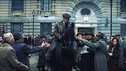Man in vintage clothing rides a black horse through a crowd in front of an old stone building; the atmosphere is tense and dramatic, with people reaching out and reacting emotionally to his presence.