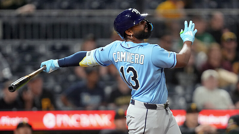 Baseball player in light blue jersey and helmet swings bat during a game, with stadium seats and spectators blurred in the background under stadium lights.