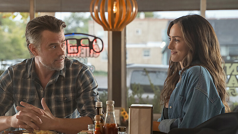 A man and woman sit at a table in a diner, facing each other and smiling, with condiments and food on the table, soft daylight coming through the windows and a cozy atmosphere in the background.