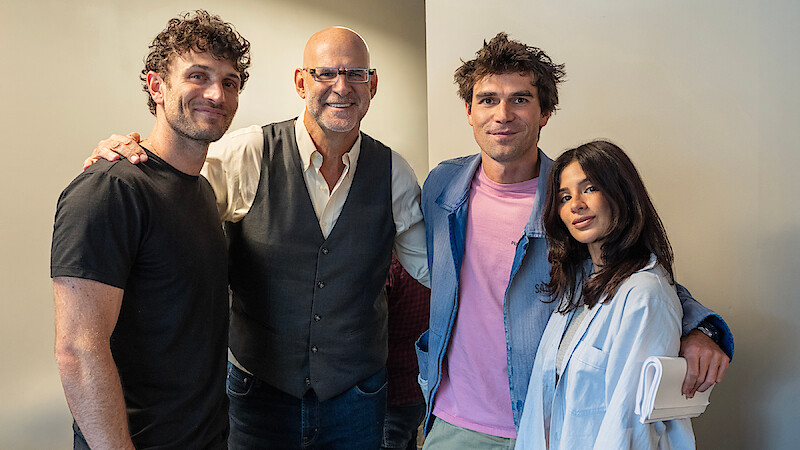 Four people standing together indoors by a beige wall, smiling at the camera. The group includes three men and one woman, casually dressed, with one person wearing glasses and another holding a rolled paper. Warm, friendly atmosphere.