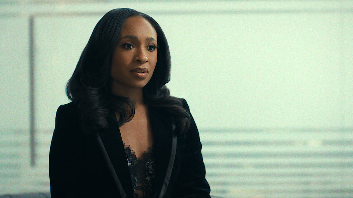 Woman with long dark hair in a black blazer sits indoors with a blurred glass wall and horizontal lines in the background, suggesting a modern office or professional setting.