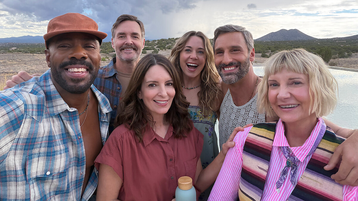 Group of six smiling adults taking a selfie outdoors by a pool, with mountains and cloudy sky in the background, casual clothing, relaxed atmosphere, desert landscape.