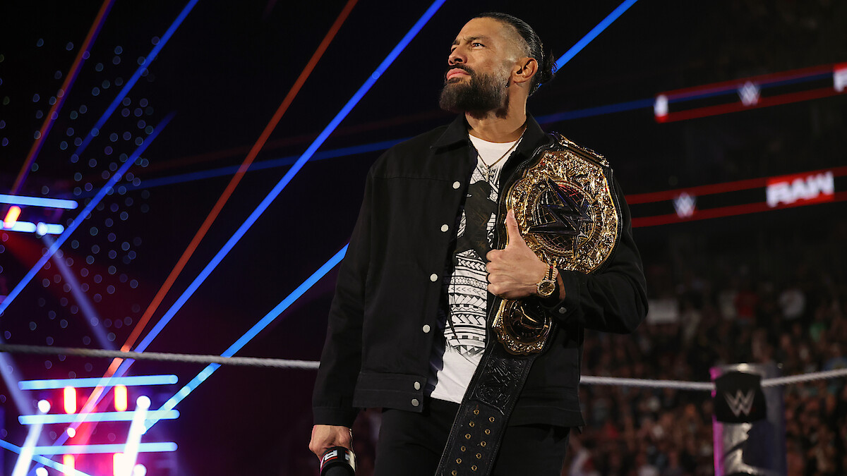 Roman Reigns holding a championship belt stands confidently in a wrestling ring, surrounded by colorful lights and a cheering crowd during a live event.