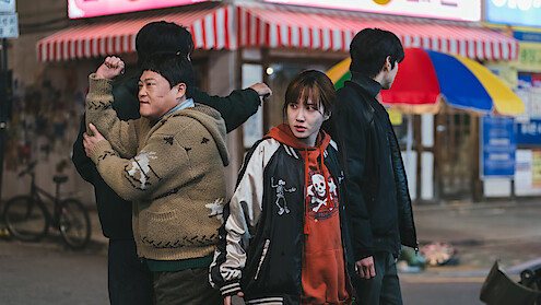 Four people stand on a city street at night near shops with striped awnings; one person looks concerned, two face away, and the street is dimly lit with visible store signs and urban elements.