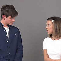 Two young people stand facing each other in front of a plain gray background, engaged in conversation. The boy wears a dark shirt and the girl wears a white top with a hair clip, both appearing serious.