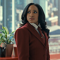 Woman in a red suit standing in a modern office with cityscape window view, looking to the side; a desk with a plant and office supplies is visible.