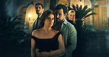 Monica Guerritore and Giacomo Gianniotti embrace in front of a coastal Italian villa. Behind them, Emanuel Caserio and Dharma Mangia Woods stand brooding.