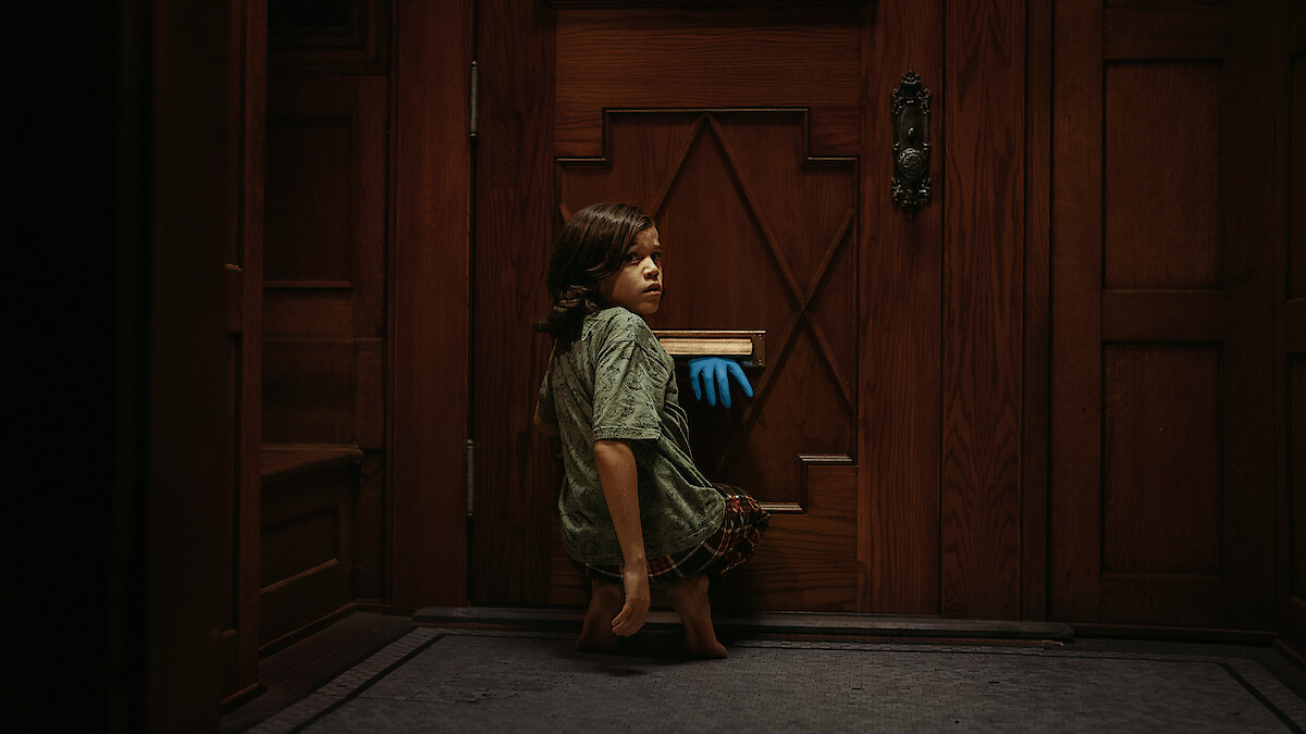 A child crouches in front of a wooden door inside a dimly lit room, looking back as a blue-gloved hand reaches through the mail slot toward them.