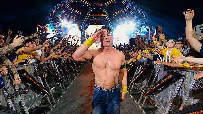 Shirtless wrestler saluting and walking down a ramp, surrounded by cheering fans at a brightly lit arena event, with colorful lights and stage setup in the background.
