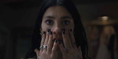 A woman with dark hair and black nail polish holds her hands to her face indoors, wearing multiple rings and looking surprised or concerned in a dimly lit setting.