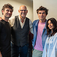 Four people posing together indoors, smiling. The group consists of three men and one woman standing closely, dressed in casual and semi-casual clothing, in a well-lit room with neutral-colored walls.