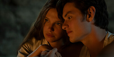 Two young people sit closely together in a dimly lit, cozy setting, with warm light illuminating their faces, suggesting an intimate or reflective moment.