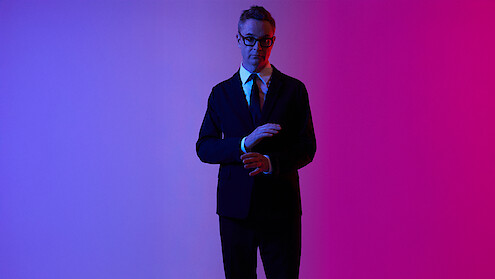 Nicolas Winding Refn wears a black suit, white shirt, and dark tie, and adjusts his cuffs against a pink and purple neon background. 