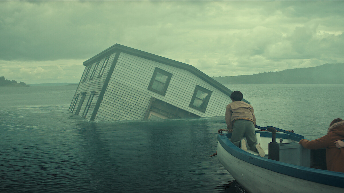Two people in a small boat approach a partially submerged house tilted in the middle of a misty lake under a cloudy sky, with forested hills in the background.