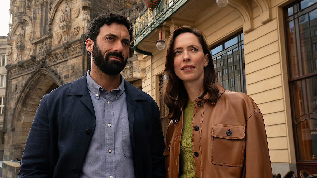 A man and woman stand outdoors in a city setting, with historic stone building details and modern architecture visible behind them. Both appear thoughtful, dressed in casual jackets, under daylight.