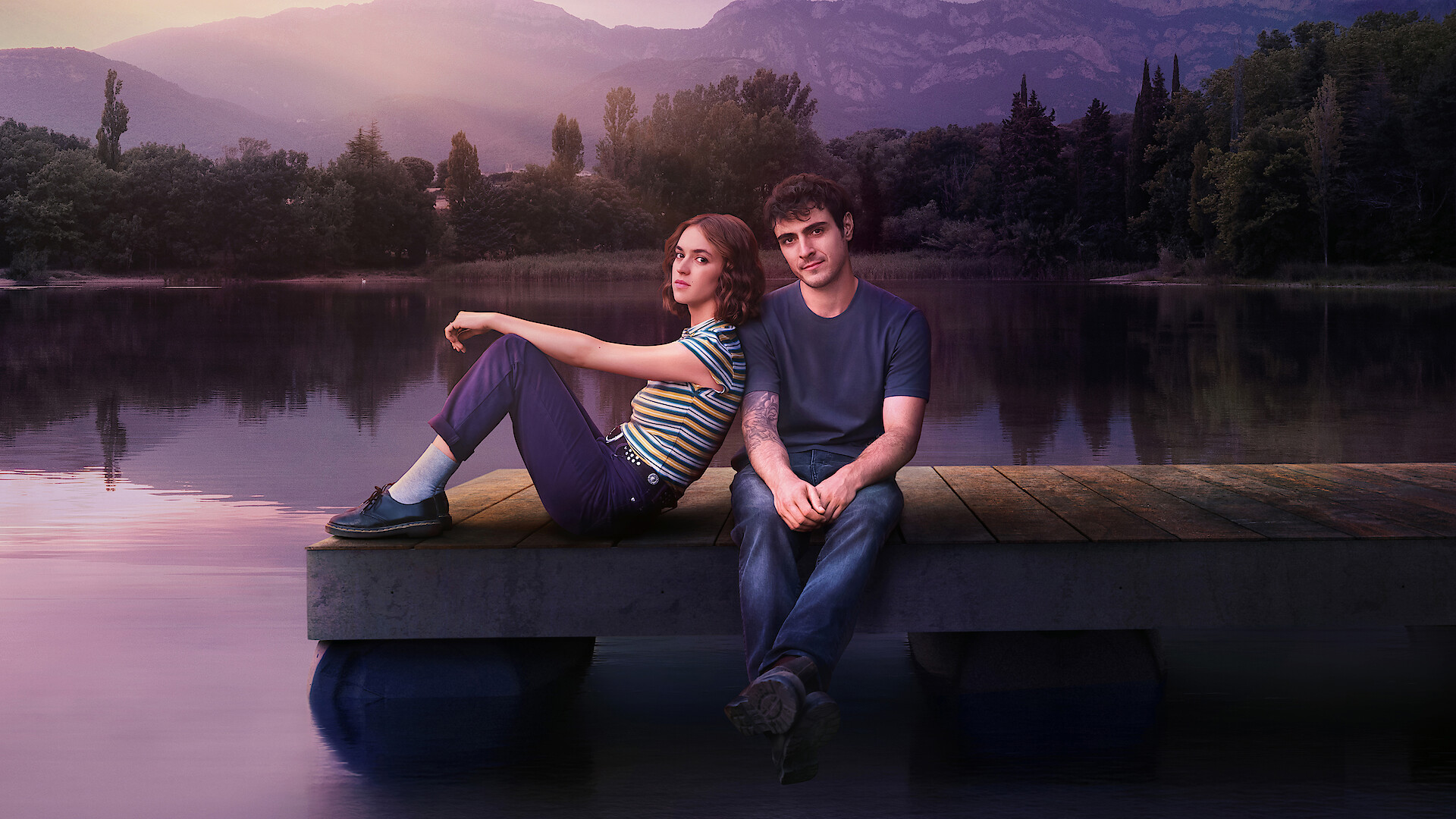 Two people sit on a wooden dock by a calm lake at sunset, surrounded by trees and mountains, with soft sunlight casting a peaceful, serene atmosphere.