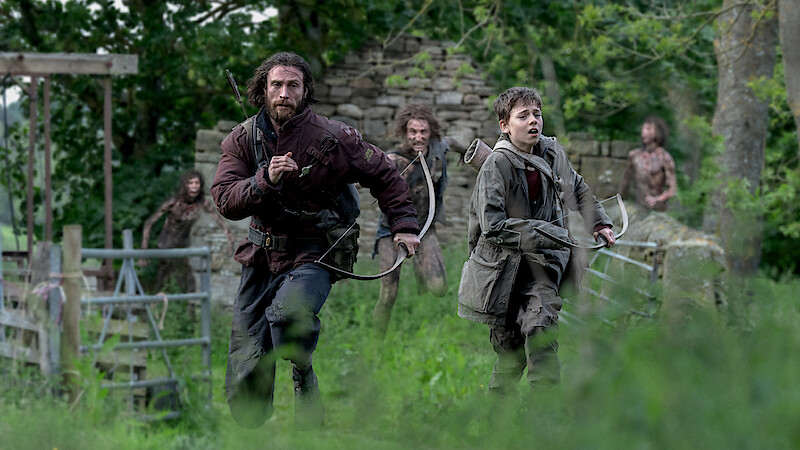 Two people, one man and one boy, run through a grassy, wooded area with stone ruins, both holding bows, while several figures follow behind them in the background. The scene is tense and outdoors in a natural environment.