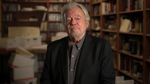 UFO reporter George Knapp sits in a library corner  facing the camera while wearing a black suit jacket over a gray collared shirt.